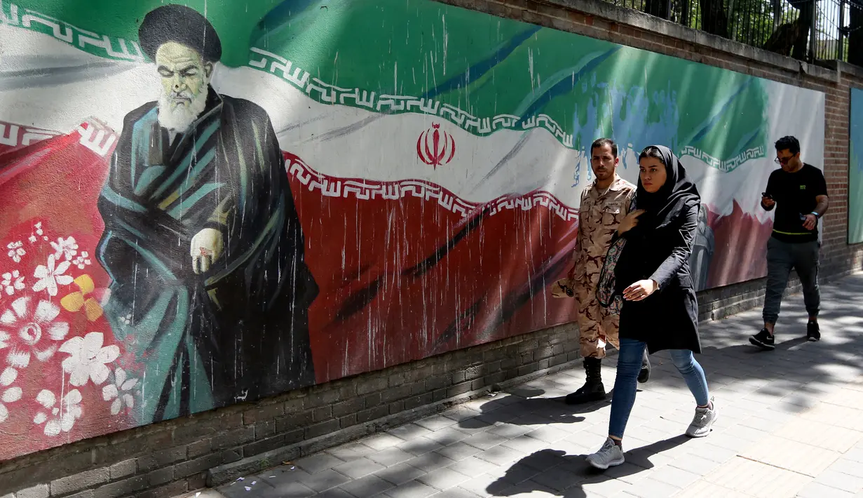 Pejalan kaki melewati lukisan mural yang menggambarkan pendiri Republik Islam Iran Ayatollah Ruhollah Khomeini dan bendera nasional di sepanjang dinding bekas Kedutaan Amerika Serikat (AS) di Ibu Kota Teheran, Iran, Sabtu (22/6/2019). (ATTA KENARE/AFP)
