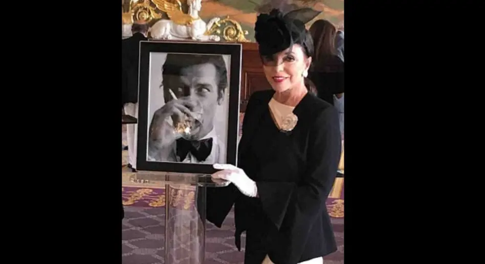 Artis senior Dame Joan Collins berpose bersama foto Roger Moore di acara pemakaman pemeran James Bond (Dailymail)