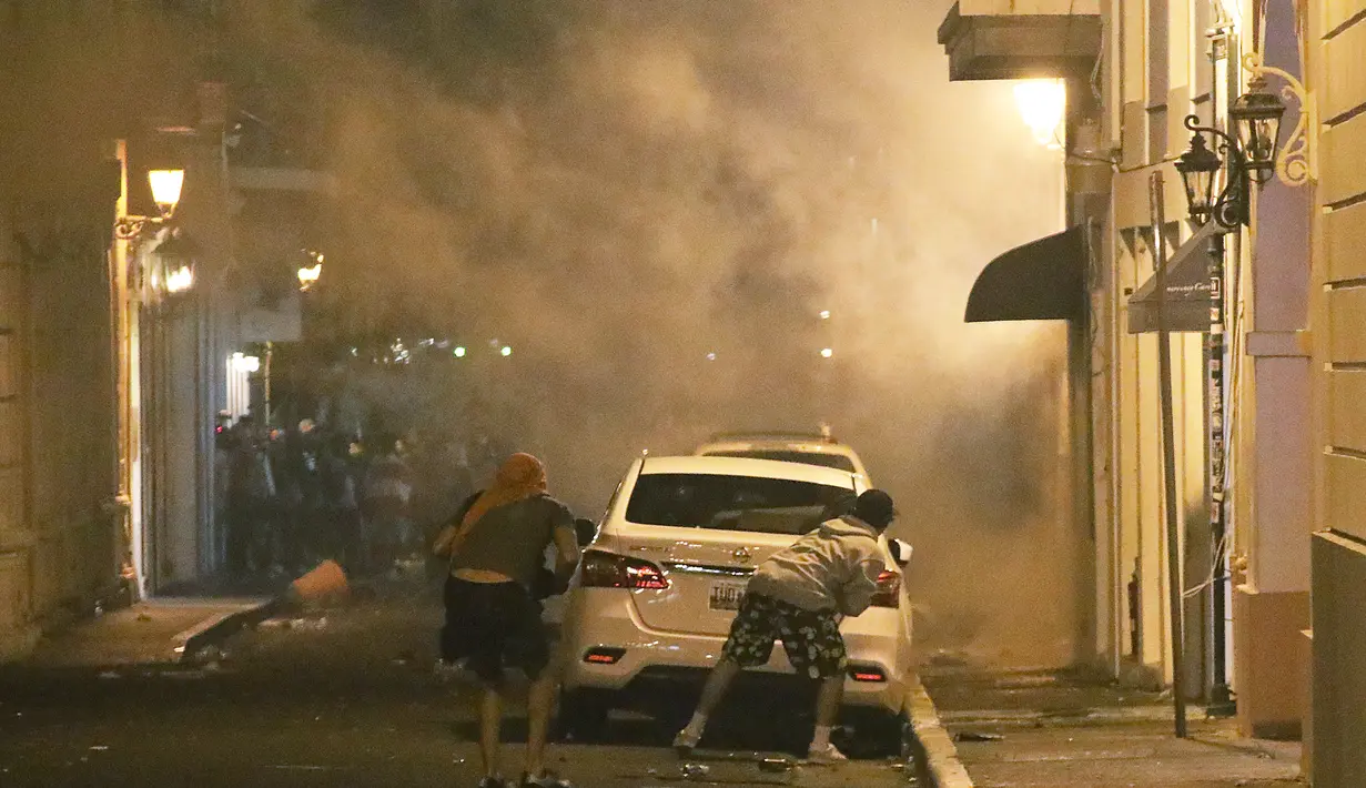 Pengunjuk rasa berlindung di belakang mobil saat bentrok dengan polisi di Old San Juan, Puerto Rico (22/7/2019). Para pengunjuk rasa menyerukan Gubernur Rosselló untuk mundur setelah obrolan kelompok terbuka termasuk komentar misoginis dan homofobik. (AFP Photo/Joe Raedle)