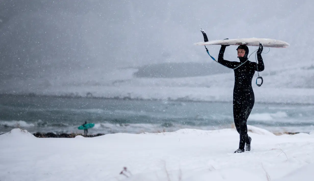 Seorang surfer wanita berjalan keluar dari air membawa papannya di bawah hujan salju di Unstad, Norwegia Utara (11/3). (AFP Photo/Olivier Morin)