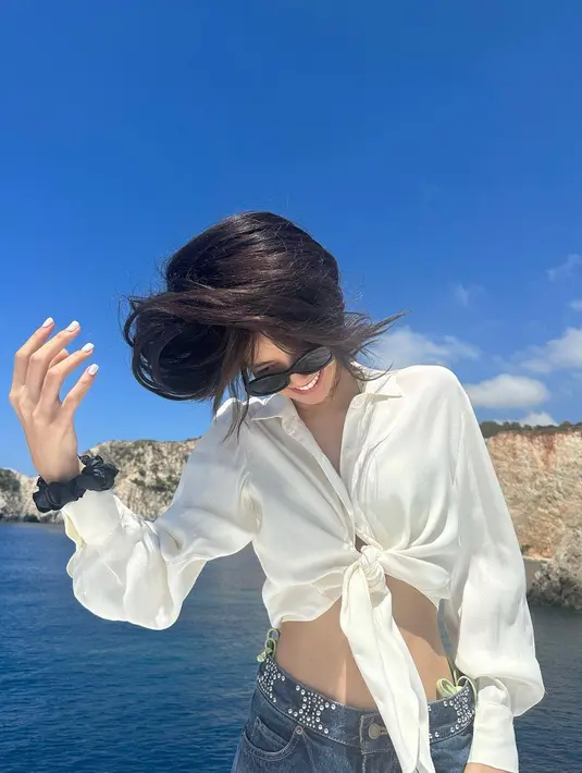 Berlatarkan tebing dan langit yang cerah, Lisa mengenakan busana kasual dengan baju putih. Dia memadukan penampilannya dengan kacamata hitam dan tersenyum cerah sambil memainkan rambut panjangnya.