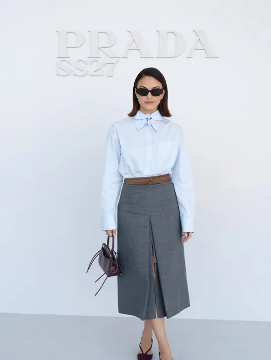 Camila Mendes mengenakan kemeja biru muda dengan tali leher pita, dipadukan rok lipit abu-abu berbelah, tas Prada marun, dan heels senada [dok. Prada]