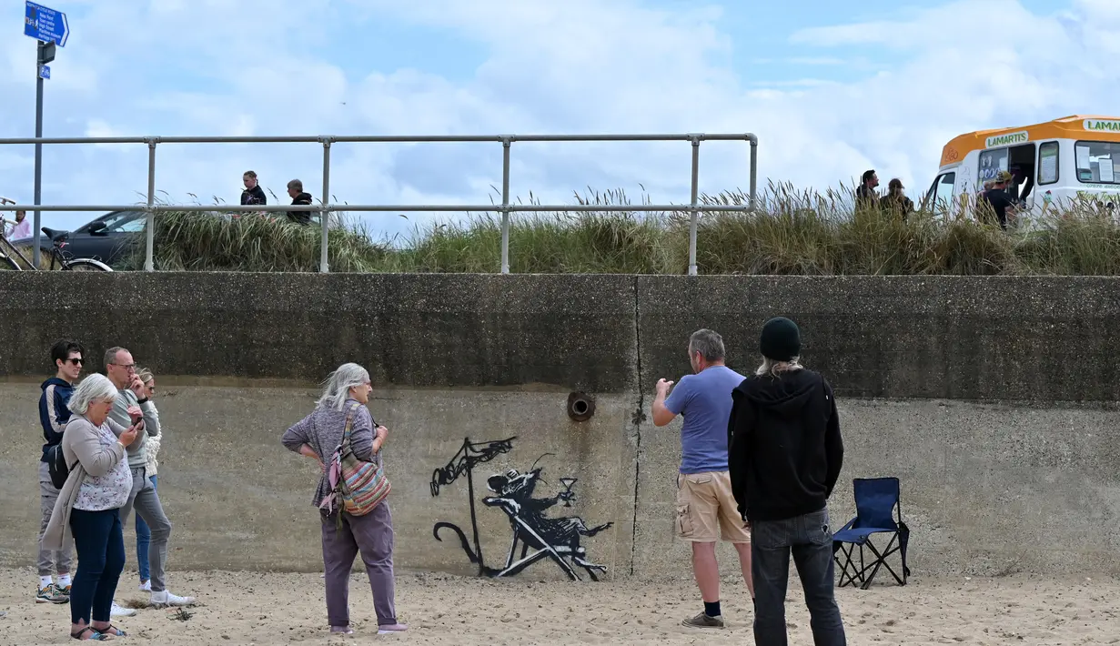 Orang-orang berkumpul untuk melihat karya seni grafiti tikus yang minum koktail, yang menyandang ciri khas seniman jalanan Banksy, di dinding di North Beach di Lowestoft di pantai Timur Inggris pada 8 Agustus 2021. Karya tersebut belum diklaim oleh Banksy. (JUSTIN TALLIS / AFP)