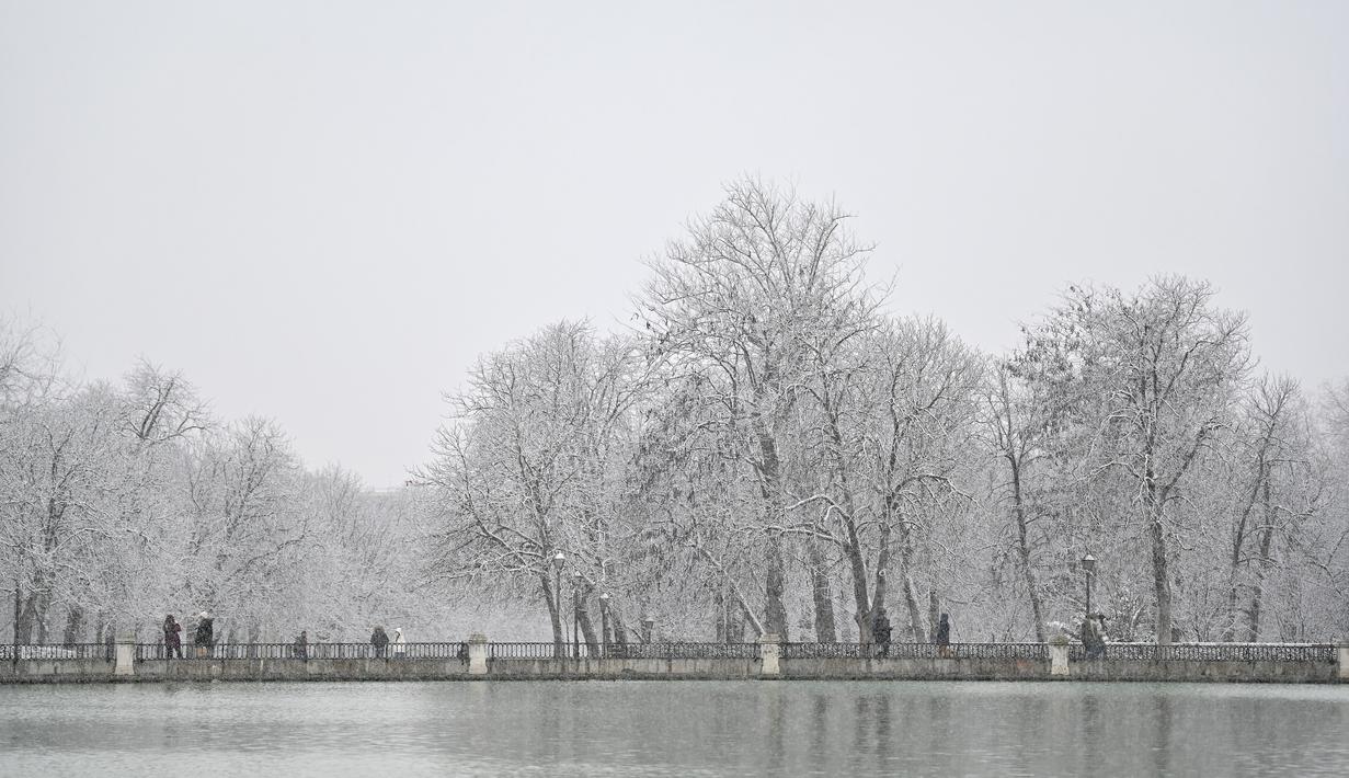 Orang-orang berjalan saat salju turun di Taman Retiro di pusat kota Madrid (7/1/2021). Pihak berwenang menyarankan warga untuk menghindari perjalanan darat di daerah dengan peringatan kondisi cuaca buruk. (AFP/Gabriel Bouys)