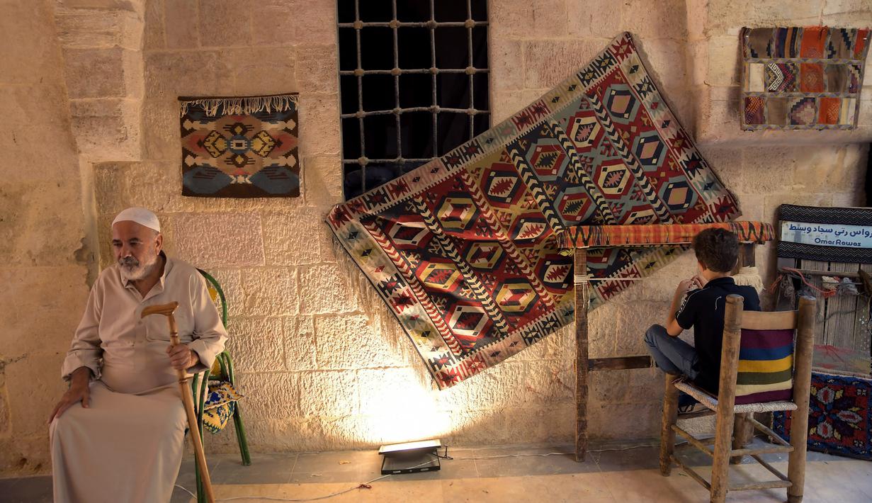 Seorang anak laki-laki bekerja di alat tenun menenun karpet tradisional sementara seorang lelaki tua duduk di sepanjang gang di "Souq Khan al-Harir" (pasar tertutup sutra) yang dibuka kembali setelah konflik bertahun-tahun di kota utara Aleppo, Suriah (29/8/2021). (AFP Photo)