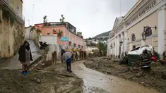 Orang-orang berjalan melalui lumpur di jalan yang banjir setelah hujan deras memicu tanah longsor yang meruntuhkan bangunan dan menyebabkan sebanyak 12 orang hilang, di Casamicciola, di pulau selatan Italia Ischia, Sabtu, 26 November 2022. Petugas pemadam kebakaran sedang melakukan upaya penyelamatan karena bala bantuan sedang dikirim dari Napoli terdekat, tetapi mengalami kesulitan dalam mencapai pulau baik dengan perahu motor atau helikopter karena cuaca. (AP Photo/Salvatore Laporta)