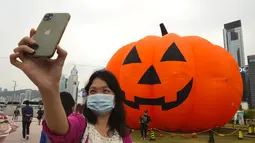 Seorang wanita mengenakan masker untuk mencegah penyebaran virus corona, berpose untuk selfie di depan dekorasi buah labu besar untuk Halloween di sebuah taman di Hong Kong, Rabu (27/10/2021). Tahun ini, perayaan tersebut jatuh pada hari Minggu, 31 Oktober 2021. (AP Photo/Vincent Yu)