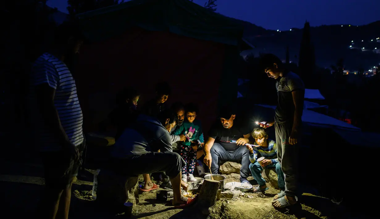 Para imigran menyiapkan makanan mereka di dekat sebuah kamp pengungsi di pulau Samos, Yunani (13/11/2019). Ribuan imigran berjuang untuk menemukan ruang di perbukitan di atas kota, membuat rumah sementara dari bahan bangunan apa pun yang dapat mereka temukan. (AFP Photo/Angelos Tzortzinis)