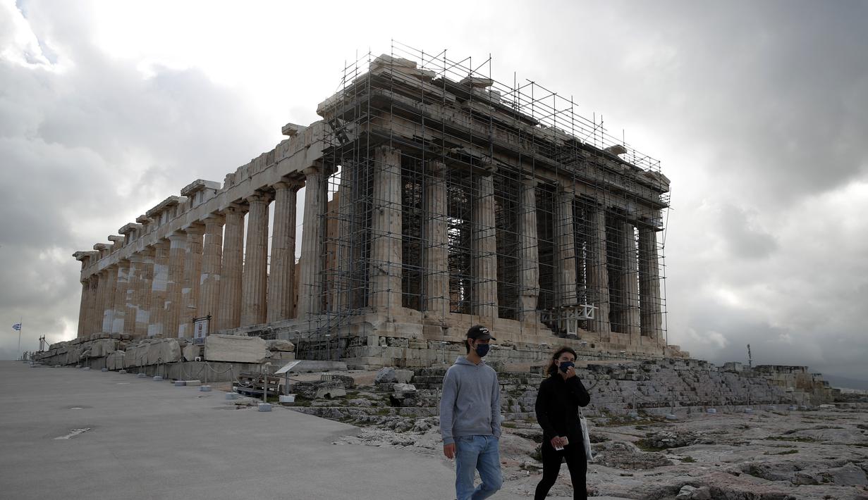 Pengunjung pertama yang mengenakan masker wberjalan di atas bukit Acropolis, dengan latar belakang kuil Parthenon di Athena, Senin (22/3/2021). Yunani membuka kembali Acropolis dan situs kuno lainnya secara nasional untuk memulai kembali musim pariwisata pada pertengahan Mei. (AP Photo/Th