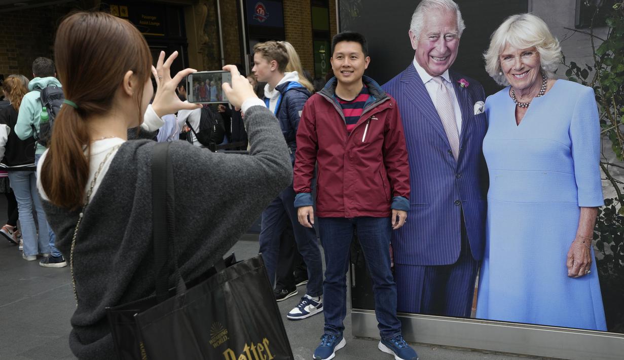 Seorang turis berpose di samping potret besar Raja Charles III dan Ratu Camilla  yang dipajang di Stasiun Kings Cross di London, Senin, 1 Mei 2023. Penobatan Raja Charles III akan berlangsung di Westminster Abbey pada 6 Mei. (AP Photo/Kirsty Wigglesworth)