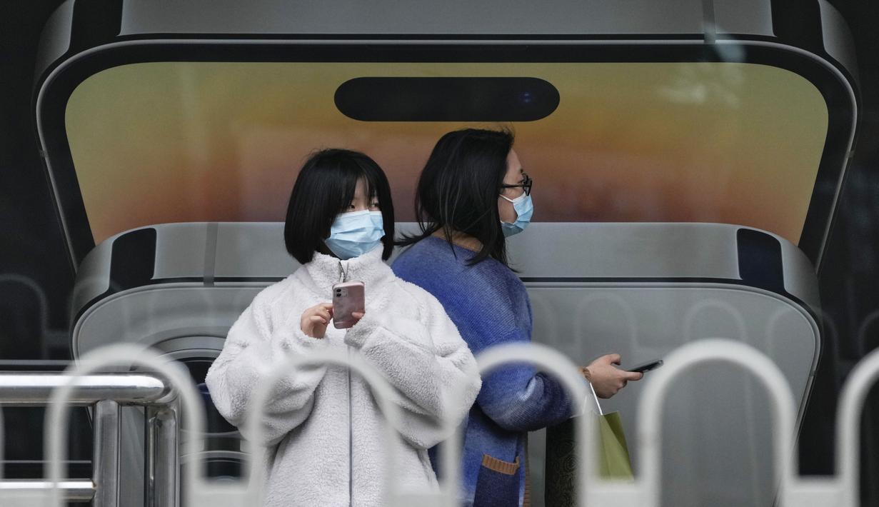 Seorang wanita yang mengenakan masker berjalan dengan yang lain menggunakan iPhone saat mereka menunggu bus mereka di halte bus yang menampilkan iklan iPhone di Beijing pada Minggu (30/10/2022). Para pekerja iPhone Apple Inc meninggalkan pabrik karena lokasinya berada dalam zona industri Kota Zhengzhou yang sedang diberlakukan lockdown setelah adanya 64 laporan kasus virus corona di kawasan tersebut. (AP Photo/Andy Wong)