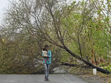 Seorang pria menggendong anak saat berjalan melewati pohon yang tumbang akibat terjangan siklon Tauktae di dekat Amreli, India, Selasa (18/5/2021). Topan Tauktae disebut yang terbesar melanda wilayah itu dalam beberapa dekade terakhir. (Punit PARANJPE / AFP)