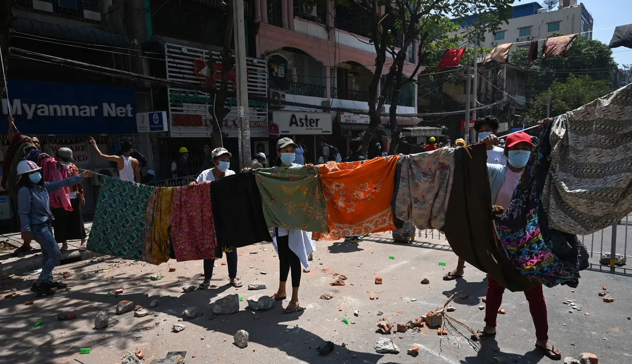 Perempuan menggantung pakaian tradisional Myanmar, longyi, di seberang jalan selama demonstrasi menentang kudeta militer di Yangon, Senin (8/3/2021). Jemuran kain itu untuk membatasi gerak polisi dan tentara karena berjalan di bawah jemuran pakaian ini dianggap akan membawa sial bagi pria. (STR/AFP)