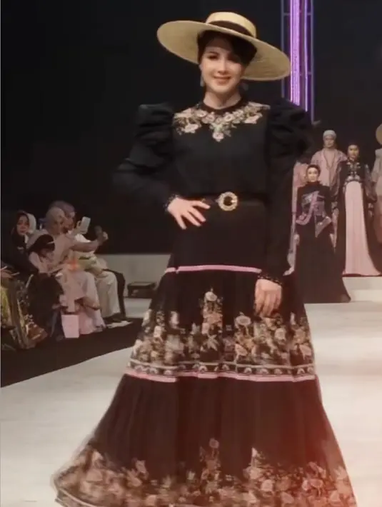 Arumi melangkah dengan percaya diri di hadapan para tamu yang hadir, memakai dress panjang berwarna hitam dengan aksen bordiran bunga yang apik di panel bagian depan.