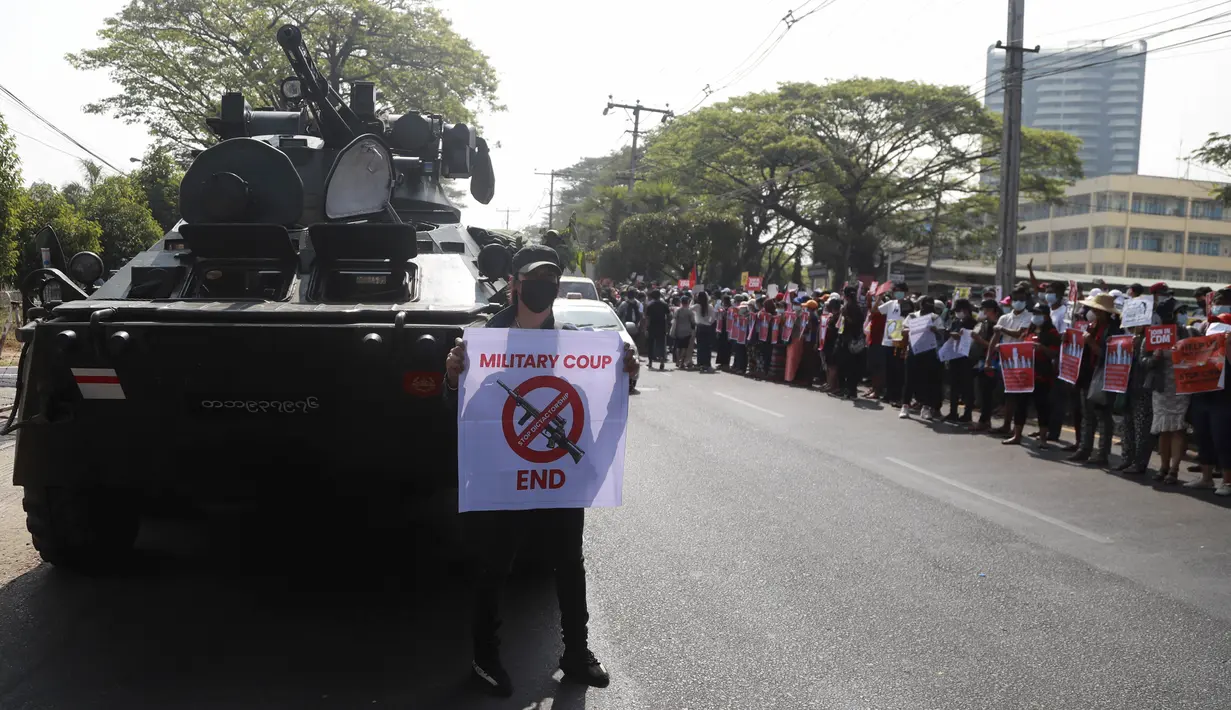 Seorang pengunjuk rasa anti-kudeta dengan poster berdiri di depan kendaraan lapis baja yang dikerahkan di luar gedung Bank Sentral Myanmar di Yangon, Senin (15/2/2021). Pasukan militer Myanmar mulai menerjunkan kendaraan lapis baja di kota-kota besar di Myanmar dan memutus akses internet. (AP Photo)