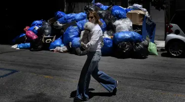 Seorang wanita menggendong anjingnya berjalan melewati tumpukan sampah di pusat kota Athena, Kamis (22/6). Tumpukan sampah tampak menggunung di beberapa kota di Yunani setelah ribuan petugas pengumpul sampah melakukan mogok kerja. (ARIS MESSINIS/AFP)
