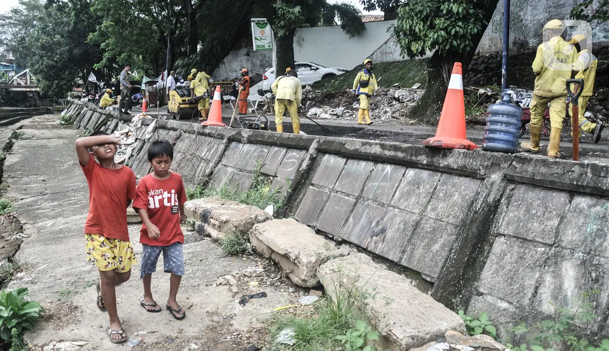 Anak-anak melintas di dekat jalan ambles yang sedang diperbaiki di Jalan Inspeksi Kali Ciliwung, Kenari, Rabu (19/1/2022). Jalan yang berada di belakang RSCM tersebut ambles sepanjang 40 meter akibat terkikis oleh hujan deras kemarin sehingga menyulitkan akses warga. (merdeka.com/Iqbal S Nugroho)