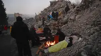 Beberapa orang duduk di dekat api unggun menunggu kabar tentang kerabat mereka di dekat puing-puing bangunan yang runtuh di Kahramanmaras, Turki, Minggu (12/2/2023). Jumlah korban tewas akibat gempa bumi dahsyat yang melanda Turki dan Suriah meningkat di atas 35.000 pada 13 Februari 2023. (OZAN KOSE/AFP)