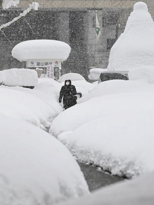 Seorang wanita berjalan di atas tumpukan salju di Toyama, di pesisir Laut Jepang, Senin (11/1/2021). Banyak yang tewas saat membersihkan salju ketika sistem udara dingin membuang hujan salju lebat di wilayah utara dan tengah negara itu. (Kyodo News via AP)