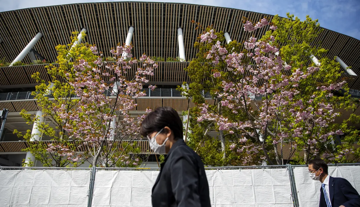 Pejalan kaki mengenakan masker melewati Stadion Nasional Jepang, tempat upacara pembukaan dan penutupan untuk Olimpiade Tokyo 2020 akan diadakan, saat bunga sakura bermekaran di atas di Tokyo (6/4/2021). Ibukota Jepang mengonfirmasi lebih dari 390 kasus virus korona baru pada hari Selasa. (AP Photo/