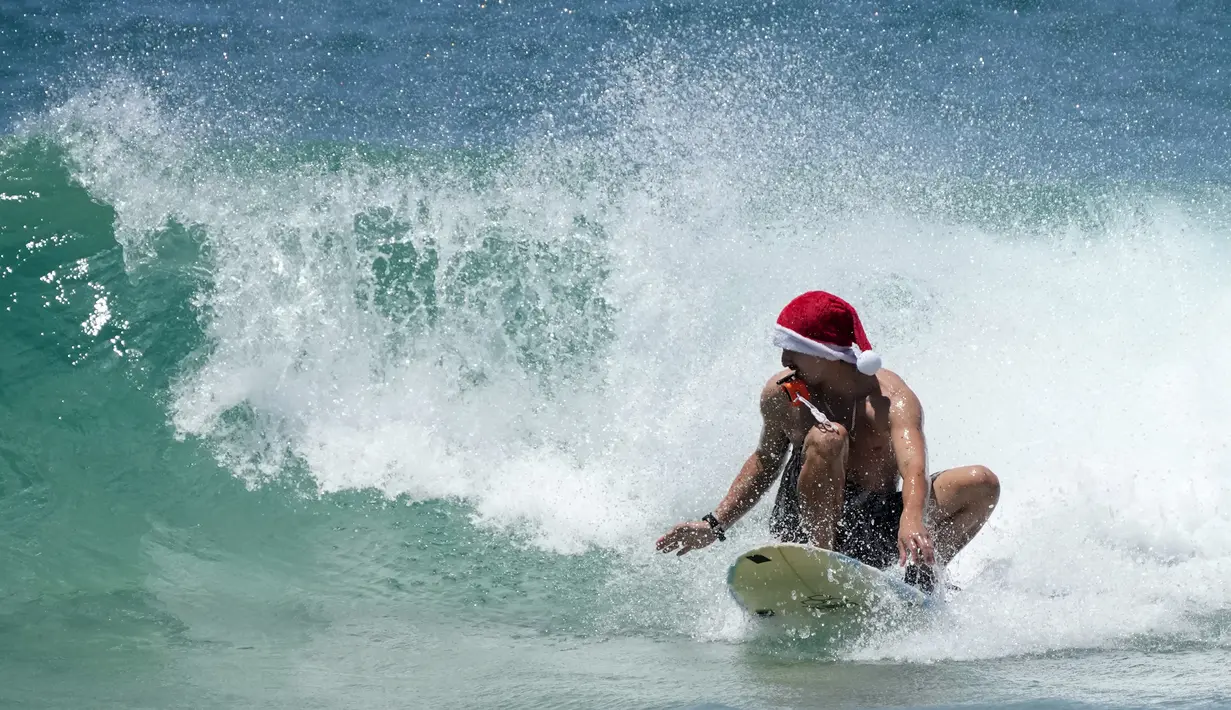 Seorang pria bertopi Santa memegang kamera aksi di giginya saat berselancar saat merayakan Natal di Pantai Bondi di Sydney, Sabtu (25/12/2021). (AP Photo/Rick Rycroft)