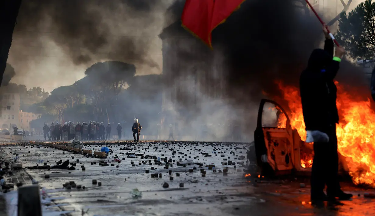 Seorang demonstran mengibarkan bendera Albania di depan mobil yang terbakar saat aksi unjuk rasa antipemerintah di luar kantor Perdana Menteri di Tirana, Albania, Sabtu (17/10/2026). (AP Photo/Hameraldi Agolli)