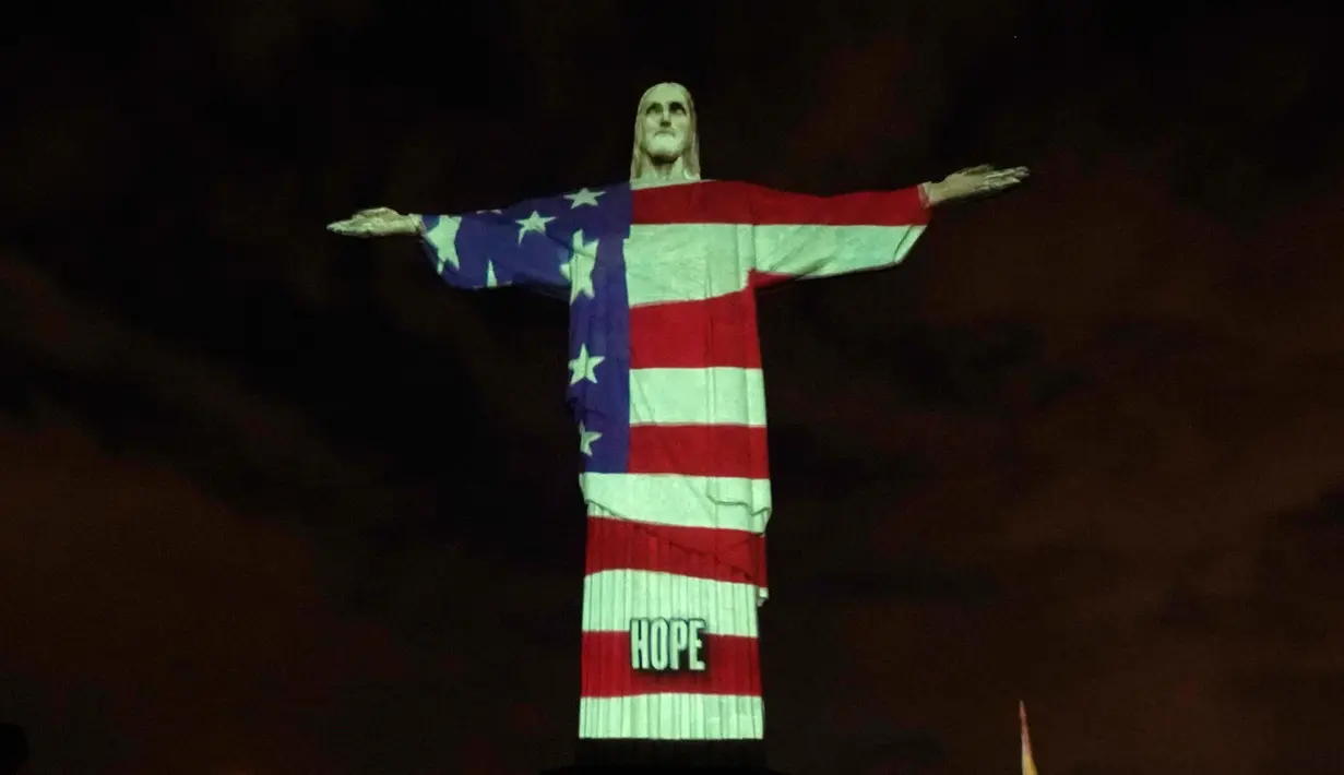 Patung Christ the Redeemer atau patung Yesus sang Penebus yang populer di dunia menyala dengan gambar bendera Amerika Serikat selama pandemi corona Covid-19 di Rio de Janeiro, Brasil, Minggu (12/4/2020). Tubuh patung itu dipenuhi gambar bendera, sekaligus pesan dan harapan. (CARL DE SOUZA/AFP)