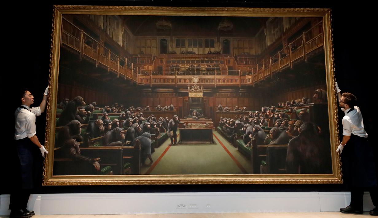 Petugas menata lukisan seniman jalanan Banksy yang dipajang di ruang lelang Sotheby, London, Jumat (27/9/2019). Lukisan dengan objek 100 simpanse yang duduk di parlemen Inggris tersebut pernah menghebohkan publik. (AP/Kirsty Wigglesworth)