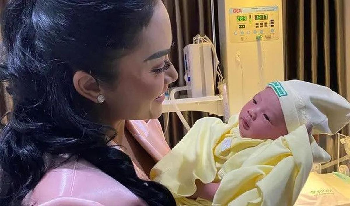 Krisdayanti memamerkan gaya saat menggendong cucu kedua Aurel-Atta dengan blazer merah muda satin.