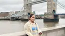 Berpose dengan latar Tower Bridge, Enzy Storia tampil modis memadukan knit dress dan coat winter warna beige. [Instagram].