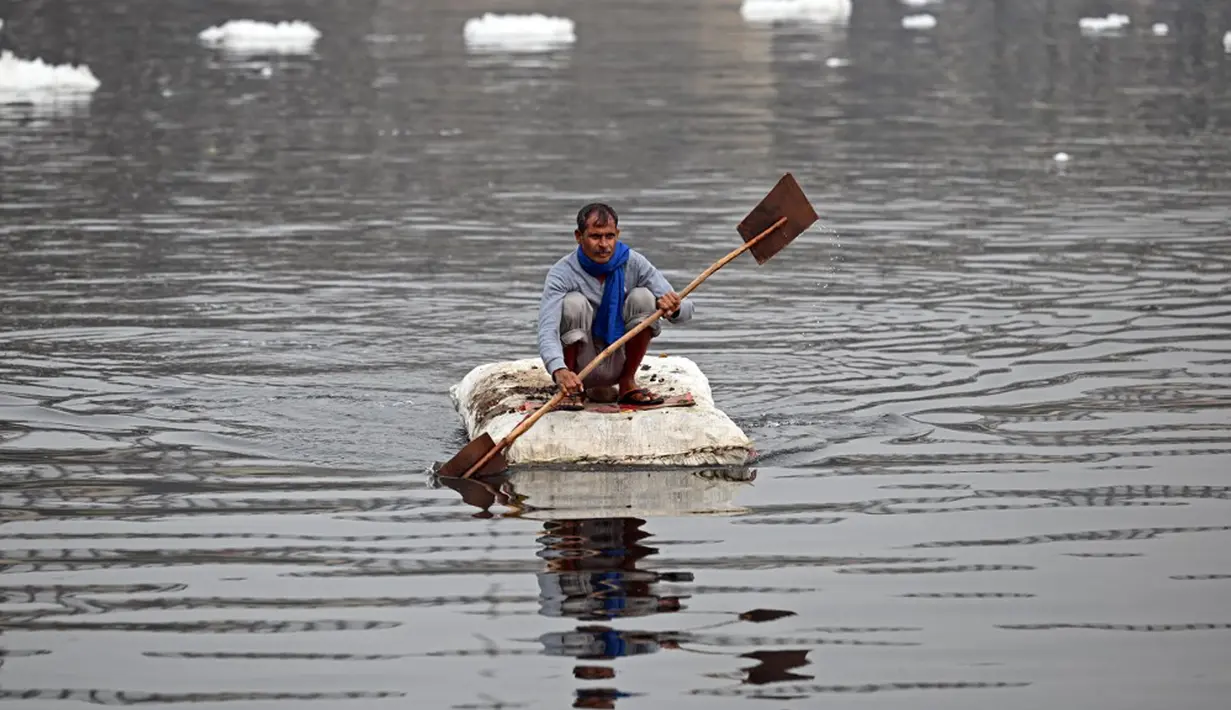 Seorang pria mendayung rakitnya sambil mengumpulkan bahan-bahan daur ulang dekat potongan busa tercemar yang mengambang di Sungai Yamuna, New Delhi, India, 26 Desember 2021. Lapisan busa beracun melapisi sungai yang dianggap suci oleh umat Hindu, Sungai Yamuna. (Sajjad HUSSAIN/AFP)