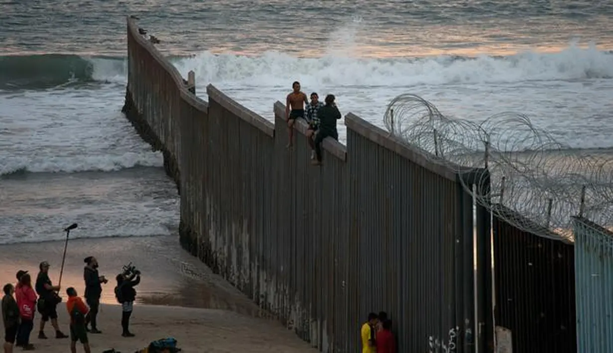 Dua imigran Honduras mengangkangi perbatasan yang memisahkan Meksiko dari AS di Tijuana, Meksiko, Rabu (21/11). Wali Kota Tijuana mendeklarasikan krisis kemanusiaan di perbatasan kotanya dengan AS. (AP Photo/Ramon Espinosa)
