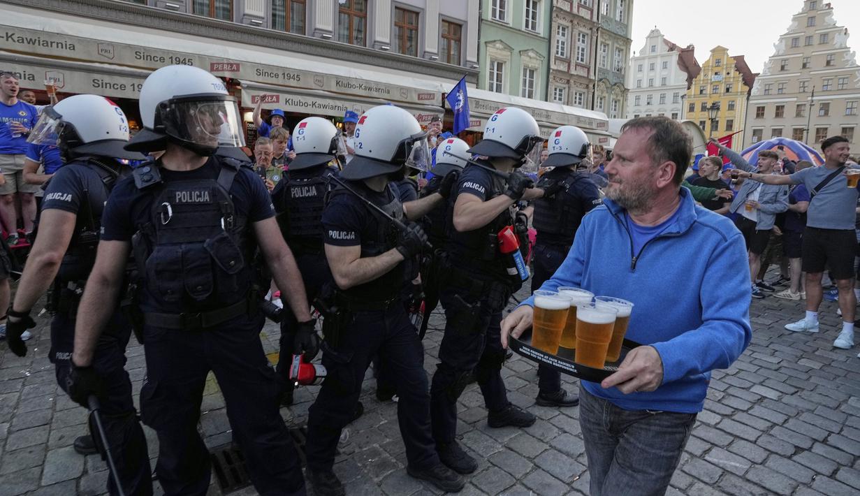 Seorang fans tengah membawa sejumlah bir melewati para polisi yang sedang membubarkan kericuhan antar fans menjelang laga final Conference League 2024/2025 antara Real Betis melawan Chelsea di sekitar restoran kota Wroclaw, Polandia, Selasa (27/05/2025) waktu setempat. (AP Photo/Czarek Sokolowski)