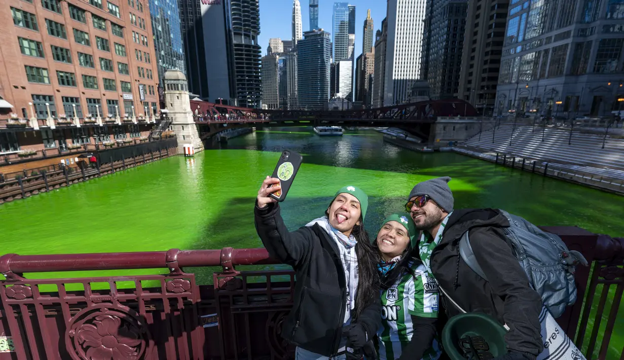 Sekelompok orang berselfie dengan latar belakang Sungai Chicago yang berwarna hijau di LaSalle Street Bridge, Sabtu (12/3/2022). Perubahan warna ini dilakukan untuk menyambut Hari St. Patrick pada Kamis mendatang. (Tyler Pasciak LaRiviere/Chicago Sun-Times via AP)