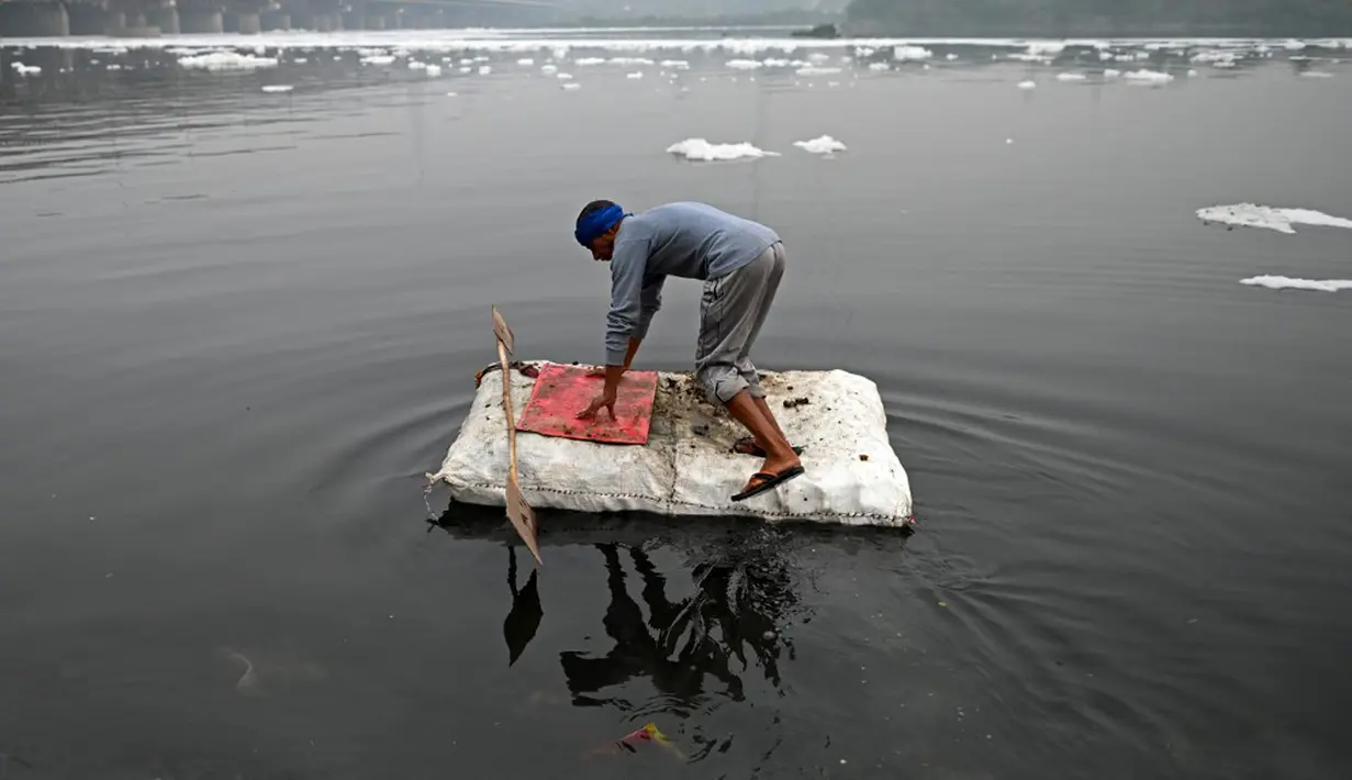 Seorang pria bersiap mendayung rakitnya untuk mengumpulkan bahan daur ulang dekat potongan busa tercemar yang mengambang di Sungai Yamuna, New Delhi, India, 26 Desember 2021. Sungai Yamuna mengalir sekitar 1.378 kilometer ke selatan dari Himalaya, melewati sejumlah negara bagian.(Sajjad HUSSAIN/AFP)