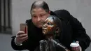 Seorang pria berswafoto dengan patung "Fearless Girl" di lokasi barunya di depan bursa efek New York, Selasa (11/12). Kini patung perunggu yang sempat menimbulkan kontroversi itu mendapatkan rumah baru yang lebih permanen. (AP/Mark Lennihan)