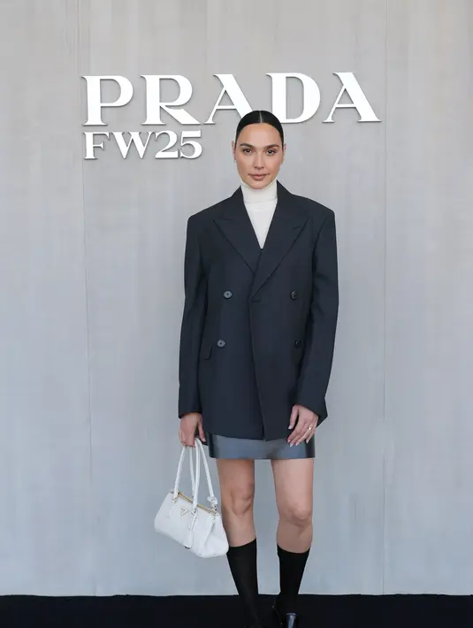 Gal Gadot tampil preppy dengan oversized jas hitam yang dipadukan turtleneck putih dan mini skirt abu-abu. Penampilannya dilengkapi dengan sepatu pantofel Prada hitam. [Dok/Prada]