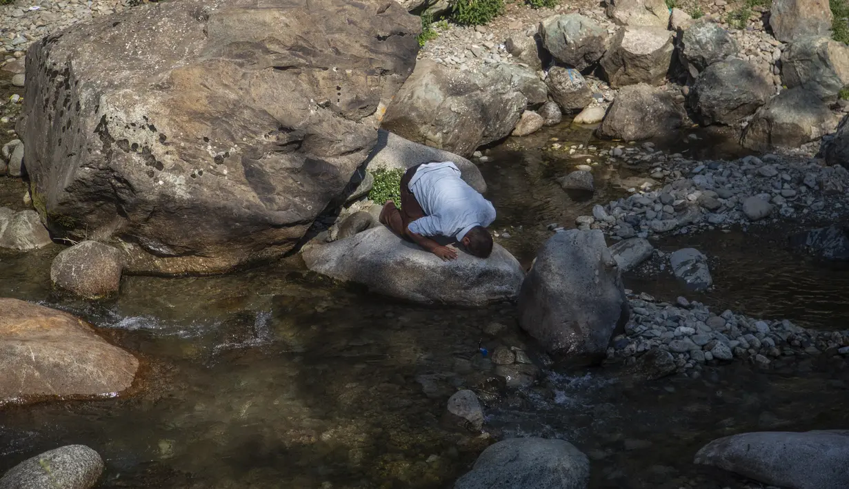 Seorang pria Kashmir yang datang dengan anak-anaknya untuk mendinginkan diri di sore yang panas berdoa di atas batu di sungai di pinggiran Srinagar, Kashmir yang dikuasai India, Minggu (4/7/2021). (AP Photo/Mukhtar Khan)