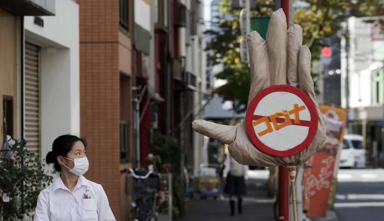 Seorang wanita mengenakan masker untuk membantu mencegah penyebaran virus corona berjalan melewati orang-orangan sawah yang menggambarkan tangan besar dengan tanda berhenti virus Corona di Tokyo, Senin (28/9/2020). Jepang mengonfirmasi lebih dari 70 kasus virus corona. (AP Photo/Eugene Hoshiko)