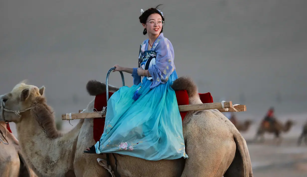 Seorang wanita mengenakan pakaian tradisional menaiki unta di padang pasir di Dunhuang di provinsi Gansu di China barat laut (10/8/2019). Sensus Tiongkok 2000 melaporkan bahwa terdapat 187,578 penduduk di kota tersebut. (AFP Photo/Str)