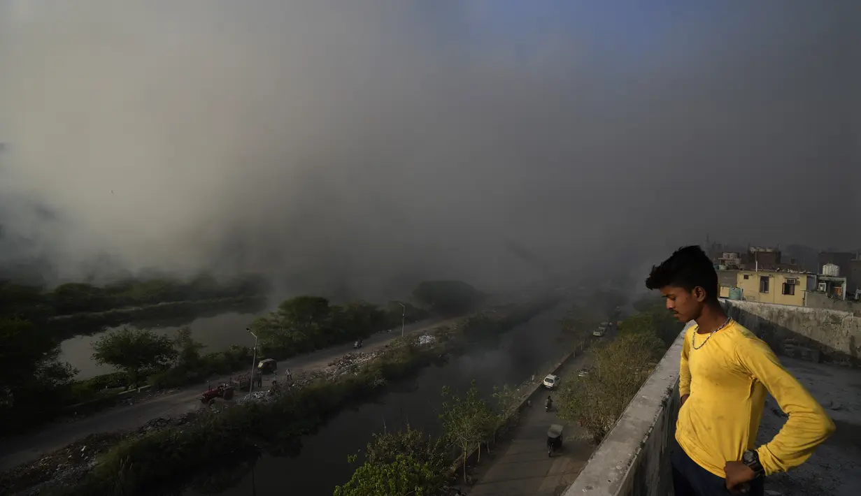 Seseorang berdiri di teras rumahnya saat api besar melalap tempat pembuangan sampah Ghazipur di New Delhi, India (28/3/2022). Kebakaran besar terjadi di tempat pembuangan sampah di daerah Ghazipur Delhi timur yang menyebabkan kepulan asap besar menyelimuti wilayah tersebut. (AP Photo/Manish Swarup)
