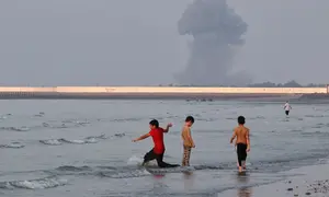Tiga anak laki-laki bermain di perairan dangkal Selat Hormuz saat kepulan asap akibat ledakan terlihat di lepas pantai Bandar Abbas, Iran, Senin (13/7/2026). (Dok. Razieh Poudat/ISNA via AP)