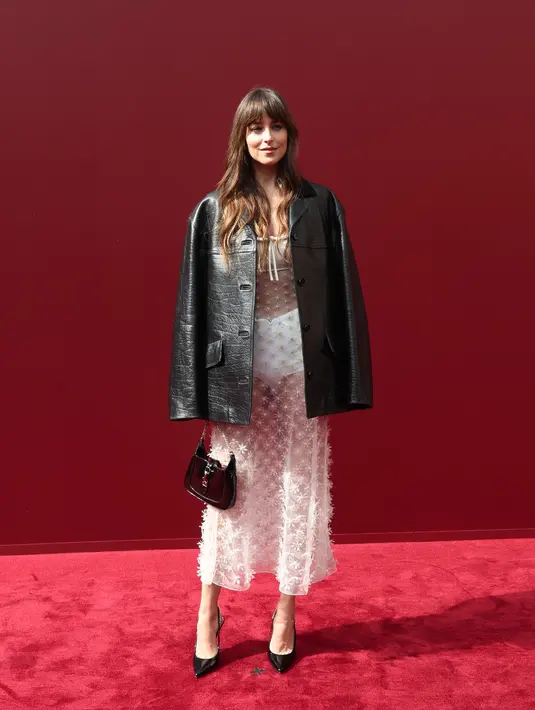 Dakota Johnson memukau dengan perpaduan sensual dan penuh gaya. Dakota Johnson mengenakan gaun putih bersulam bunga yang cukup tipis memperlihatkan dalamannya. Ia melapisinya dengan jaket kulit hitam, sepatu hak slingback paten, dan tas mini klasik jackie.  [Dok/Gucci].