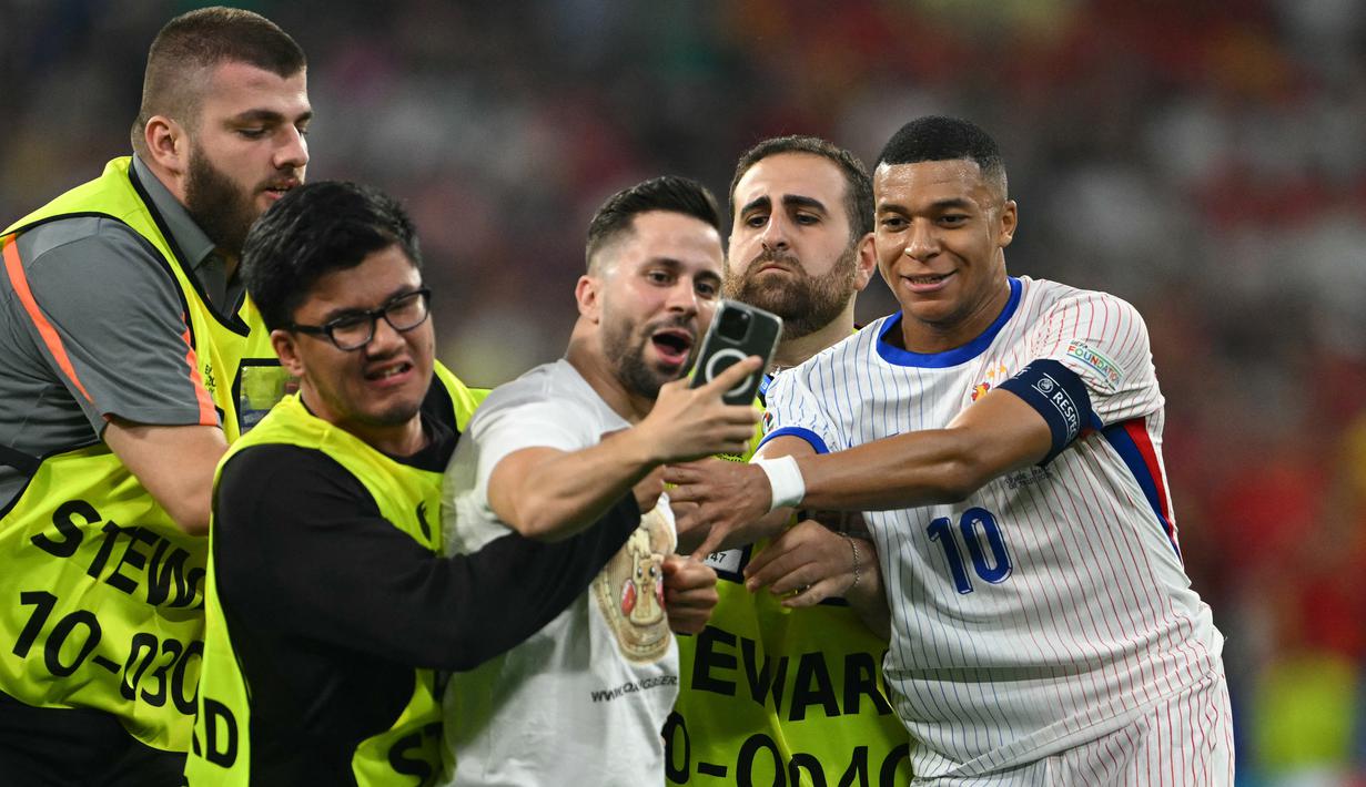 Petugas keamanan menghentikan penyerbu lapangan saat ia mengambil swafoto dengan Kylian Mbappe pada semifinal Euro 2024 antara Spanyol vs Prancis di Allianz Arena, Rabu (10/7/2024) dini hari WIB. (Fabrice COFFRINI / AFP)