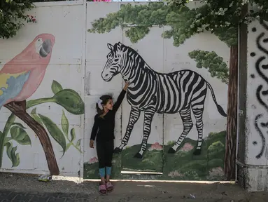 Seorang gadis Palestina menyentuh mural zebra di luar kebun binatang lokal yang ditutup di Rafah di Jalur Gaza selatan (26/5/2020). Kebun binatang itu ditutup selama liburan Idul Fitri karena pandemi virus coronavirus COVID-19. (AFP/Said Khatib)
