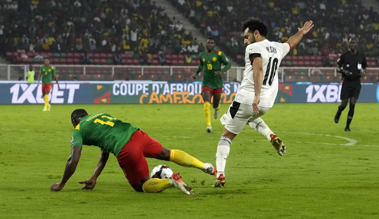 Penyerang Mesir, Mohamed Salah mencoba melakukan tendangan gawang dibayangi penyerang Kamerun Christian Bassogog pada laga semifinal Piala Afrika 2021 di Paul Biya Stadium, Jumat (4/2/2022) dini hari WIB. Mesir sukses mengalahkan Kamerun 3-1 via drama adu penalti. (AP Photo/Themba Hadebe)