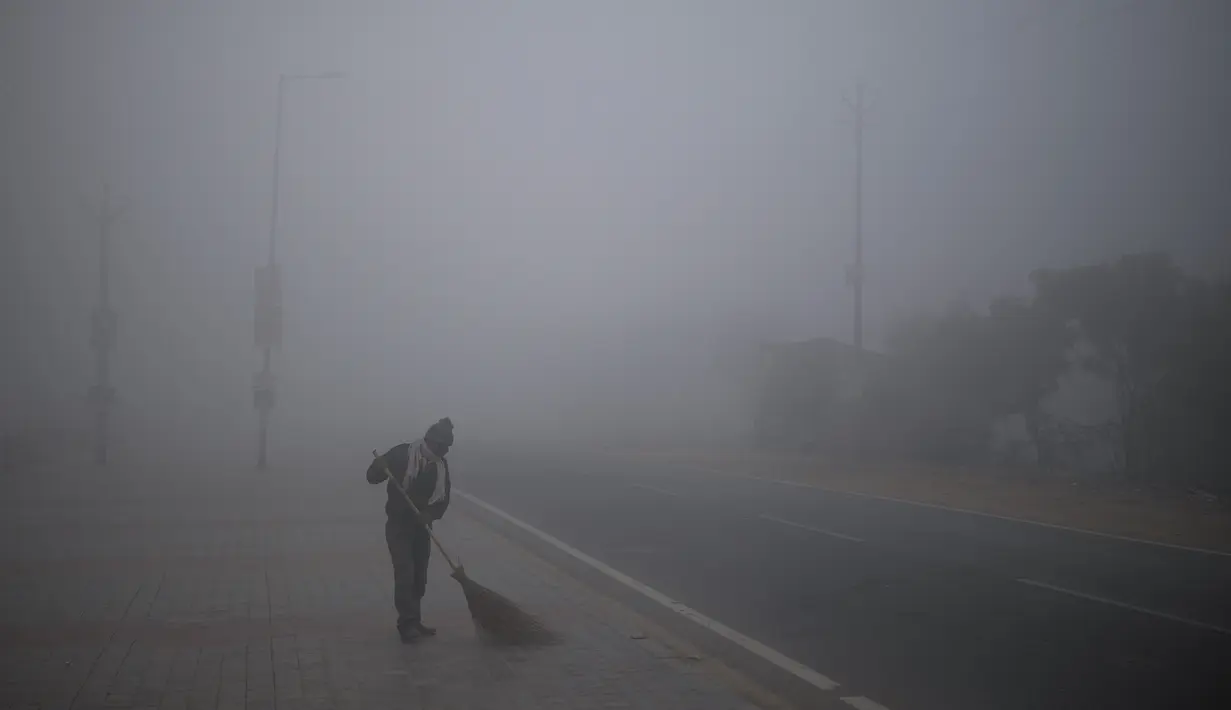 Seorang pekerja kota menyapu trotoar yang diselimuti kabut tebal pada pagi yang dingin di pinggiran New Delhi, India (1/1/2021). (AP Photo/Altaf Qadri)