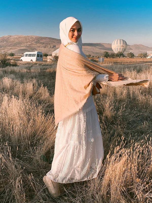 Anggunnya Citra Kirana mengenakan hijab dan dress panjang berwarna putih dipadu dengan syal krem dengan latar belakang balon udara dan padang ilalang di Cappadocia, Turki. (Liputan6.com/IG/@citraciki)