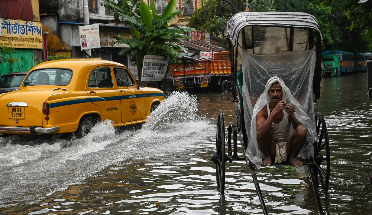 Seorang penarik becak membungkus dirinya dengan lembaran plastik sambil menunggu untuk mengangkut pelanggan melintasi jalan yang tergenang air setelah hujan lebat di Kolkata (20/9/2021). Hujan dilaporkan dipicu oleh sirkulasi siklon yang terbentuk di atas Teluk Benggala. (AFP/Dibyangshu Sarkar)