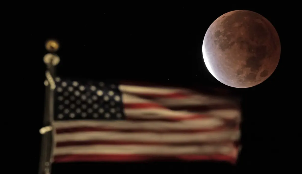 Bayangan bumi menutupi bulan purnama selama gerhana bulan sebagian atau parsial saat terbenam melewati bendera AS di atas sebuah gedung di pusat kota Kansas City, Missouri, Jumat (19/11/2021). (AP Photo/Charlie Riedel)